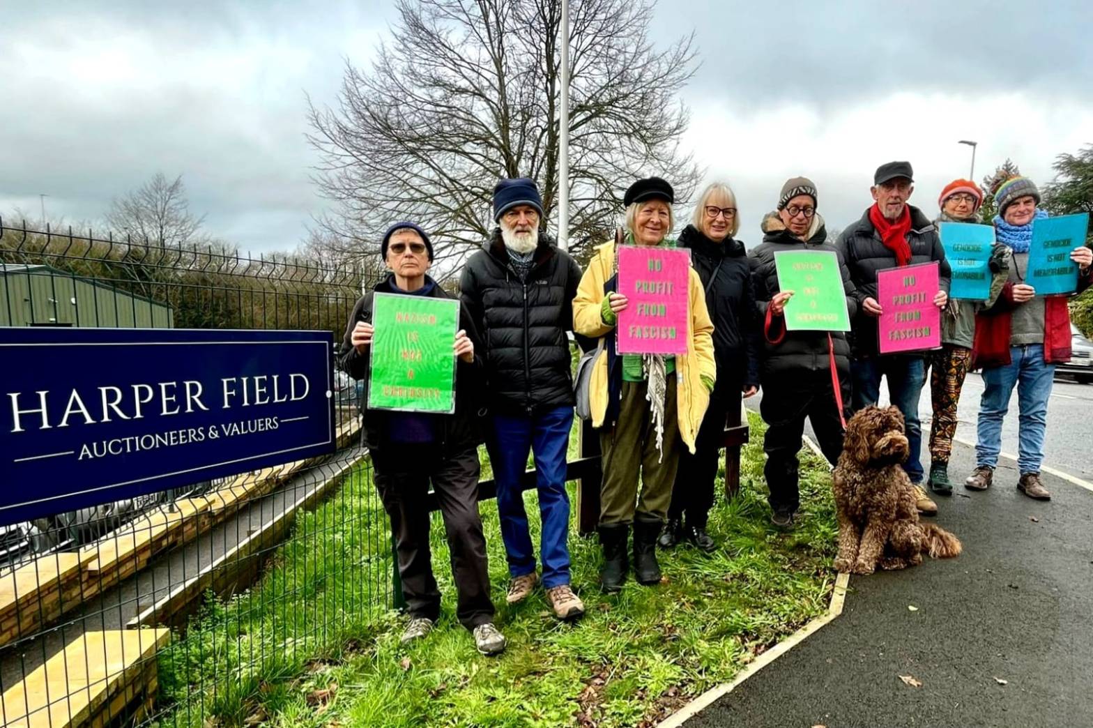Harper Field auctioneers sign and 8 people holding placards reading "Genocide is not memorabilia", "No profit from fascism", and "Nazism is not a curiosity".
